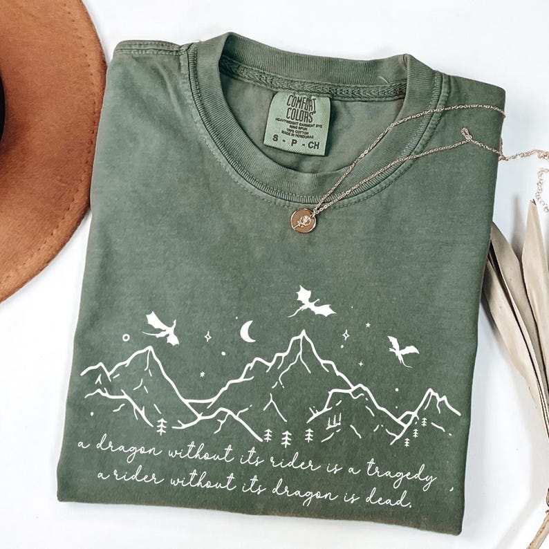 A green t-shirt with a mountain landscape design and a quote printed on it, along with a necklace and a brown hat.