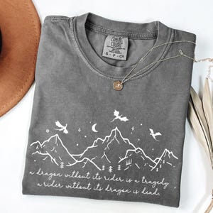 A gray t-shirt with a graphic design depicting mountains, trees, and a dragon, along with a quote that reads "A dragon without its rider is a tragedy, a rider without its dragon is dead."