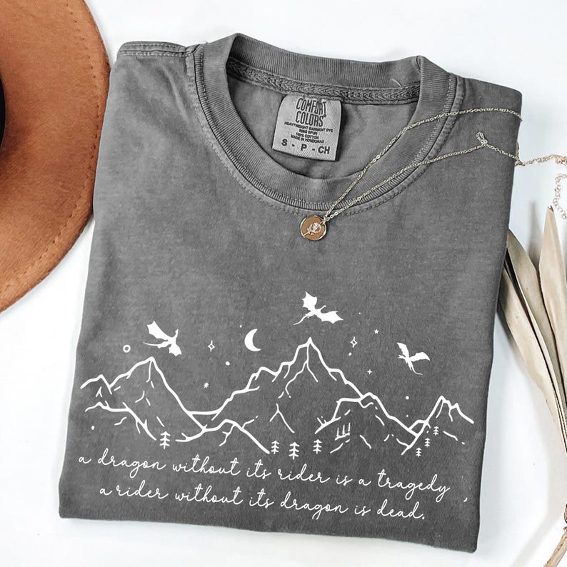 A gray t-shirt with a graphic design depicting mountains, trees, and a dragon, along with a quote that reads "A dragon without its rider is a tragedy, a rider without its dragon is dead."