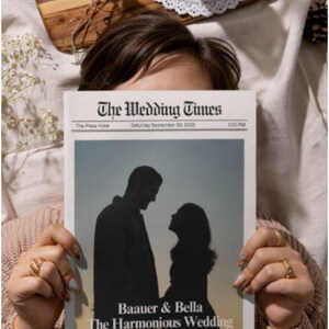 May include: A newspaper titled "The Wedding Times" featuring a silhouette of a couple. The headline reads "Baauer & Bella The Harmonious Wedding." The newspaper is held by a person wearing a sweater. A wooden tray with a succulent, glasses, and a candle is in the background.