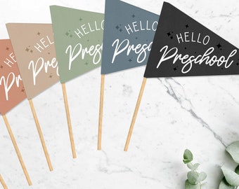 Op de afbeelding: Vijf kleurrijke driehoekige vlaggen met houten stokken, elk met de tekst "Hello Preschool" in witte letters met kleine witte sterren. De vlaggen zijn in tinten paars, oranje, beige, groen en zwart.