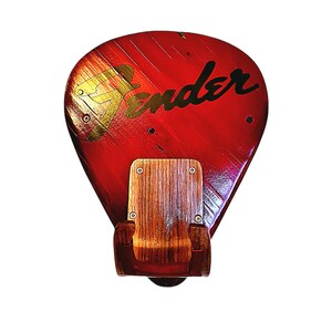 May include: A red guitar pick-shaped wall art piece with the Fender logo in black script. The wooden art has a natural wood chair attached to the bottom. The Fender logo is in gold.