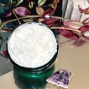 May include: A close-up of a white cream in a green jar with a purple amethyst crystal on a reflective surface.