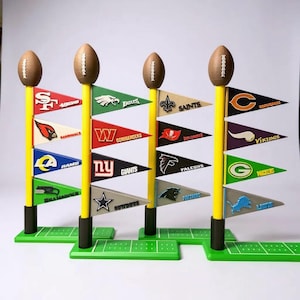 May include: A set of four miniature football field goal posts with pennant flags displaying NFL team logos. Each post has a brown football on top, yellow pole, and green base. The flags feature team names and logos.