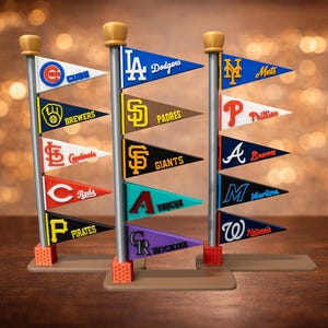 May include: Miniature pennant flags featuring various Major League Baseball team logos, including the Dodgers, Mets, and Phillies. Each flag is a different color and design, mounted on a silver pole with a gold finial. The flags are arranged on a brown base.