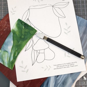 May include: A sheet with a floral design and the text "She's All Glass" is next to a glass cutter and pieces of stained glass in shades of green, blue, and red. The sheet has the text "Suggested ring placement".