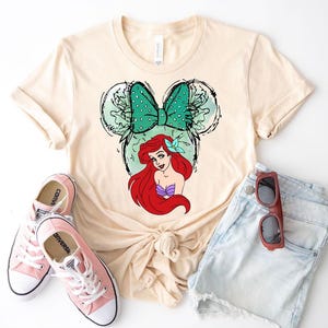 Puede incluir: Camiseta color crema con un gr&aacute;fico de Ariel de La Sirenita dentro de una silueta de Mickey Mouse. El dise&ntilde;o incluye un lazo verde azulado y un top de concha p&uacute;rpura. Zapatillas rosas y pantalones cortos de mezclilla completan el atuendo casual.