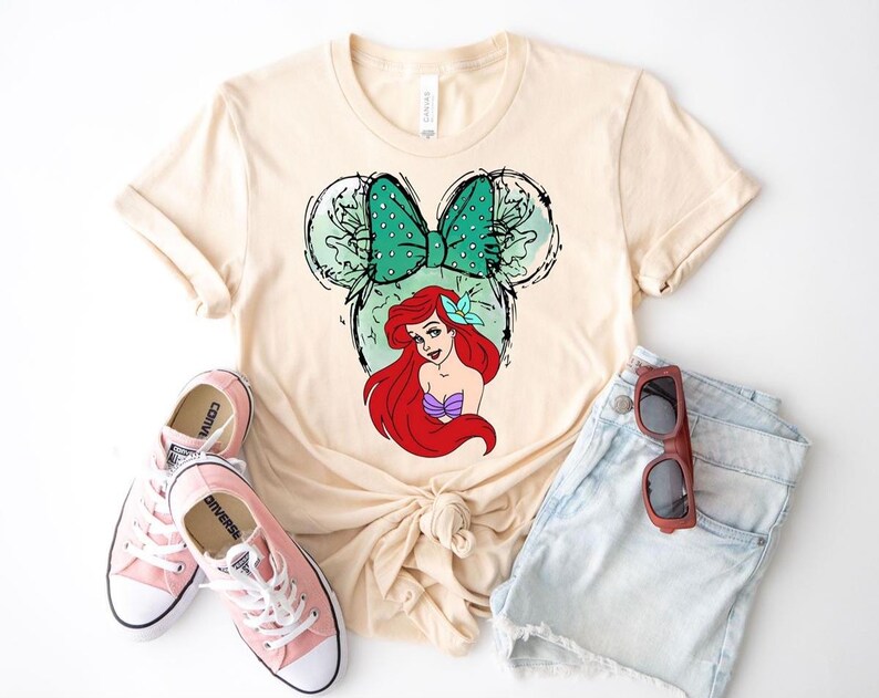 Puede incluir: Camiseta color crema con un gr&aacute;fico de Ariel de La Sirenita dentro de una silueta de Mickey Mouse. El dise&ntilde;o incluye un lazo verde azulado y un top de concha p&uacute;rpura. Zapatillas rosas y pantalones cortos de mezclilla completan el atuendo casual.
