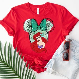 Puede incluir: Una camiseta roja con un gr&aacute;fico de Ariel de La Sirenita, dentro de un dise&ntilde;o de orejas de Mickey Mouse. El dise&ntilde;o incluye un lazo verde con lunares y el pelo rojo y el top de concha p&uacute;rpura de Ariel. La camiseta se combina con pantalones cortos de mezclilla y gafas de sol.