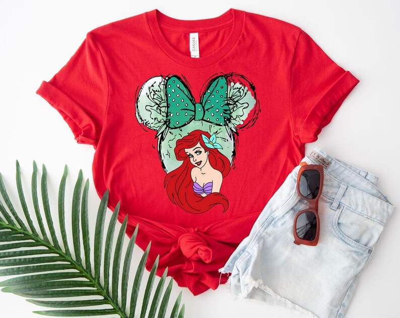 Puede incluir: Una camiseta roja con un gr&aacute;fico de Ariel de La Sirenita, dentro de un dise&ntilde;o de orejas de Mickey Mouse. El dise&ntilde;o incluye un lazo verde con lunares y el pelo rojo y el top de concha p&uacute;rpura de Ariel. La camiseta se combina con pantalones cortos de mezclilla y gafas de sol.