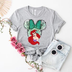 Puede incluir: Una camiseta gris con un gr&aacute;fico de Ariel de La Sirenita. El dise&ntilde;o incluye un lazo verde, orejas de rat&oacute;n y el pelo rojo y la parte superior de concha p&uacute;rpura de Ariel. La camiseta se combina con pantalones cortos de mezclilla y gafas de sol.