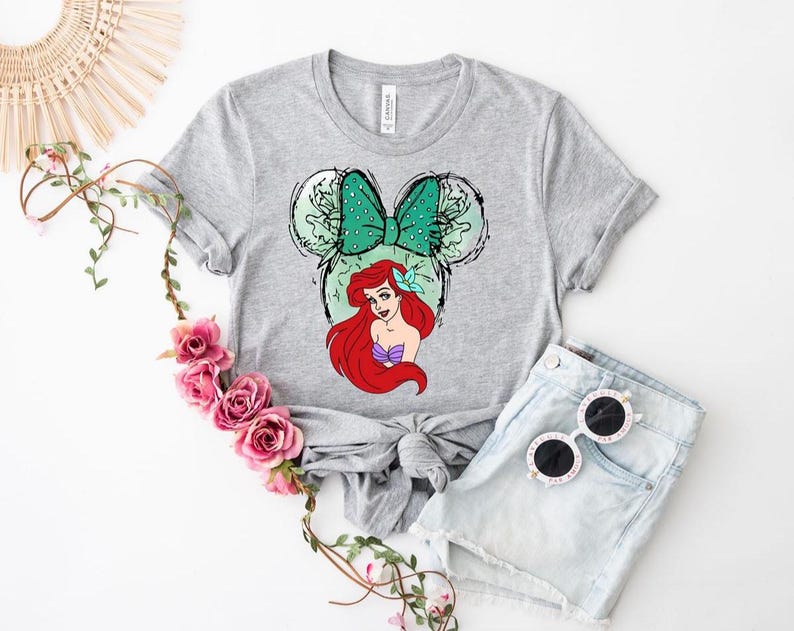 Puede incluir: Una camiseta gris con un gr&aacute;fico de Ariel de La Sirenita. El dise&ntilde;o incluye un lazo verde, orejas de rat&oacute;n y el pelo rojo y la parte superior de concha p&uacute;rpura de Ariel. La camiseta se combina con pantalones cortos de mezclilla y gafas de sol.