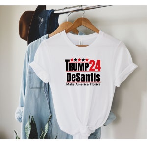 May include: A white t-shirt with the text "TRUMP 24 DeSantis Make America Florida" in black and red. The shirt is hanging on a wooden hanger. A denim shirt and a brown hat are visible in the background.
