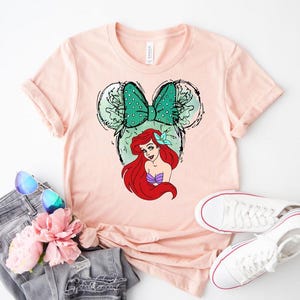 Puede incluir: Camiseta rosa claro con un dise&ntilde;o de Ariel de La Sirenita. El dise&ntilde;o incluye un lazo verde con lunares, orejas de rat&oacute;n y el pelo rojo y la parte superior de concha morada de Ariel. La camiseta se combina con vaqueros, gafas de sol y zapatillas blancas.