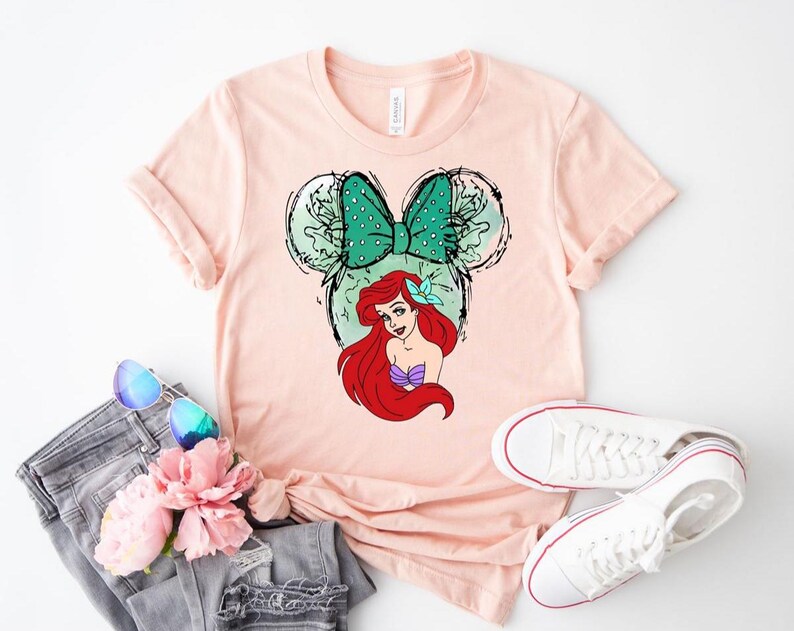 Puede incluir: Camiseta rosa claro con un dise&ntilde;o de Ariel de La Sirenita. El dise&ntilde;o incluye un lazo verde con lunares, orejas de rat&oacute;n y el pelo rojo y la parte superior de concha morada de Ariel. La camiseta se combina con vaqueros, gafas de sol y zapatillas blancas.