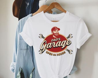 May include: A white short-sleeved t-shirt featuring a graphic print of a bearded person in a cap above a red oval. The text reads DAD'S Garage OPEN 24 HOURS, flanked by two crossed silver wrenches. The design uses red, black, and cream colors.