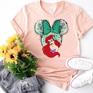 Puede incluir: Una camiseta rosa claro con un dise&ntilde;o de Ariel de La Sirenita. El dise&ntilde;o incluye un lazo verde con lunares, orejas de Mickey Mouse y el pelo rojo y la parte superior morada de Ariel. Zapatillas blancas, un ramo de flores amarillas y unos vaqueros grises tambi&eacute;n est&aacute;n en la imagen.