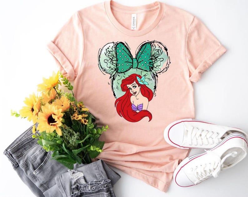 Puede incluir: Una camiseta rosa claro con un dise&ntilde;o de Ariel de La Sirenita. El dise&ntilde;o incluye un lazo verde con lunares, orejas de Mickey Mouse y el pelo rojo y la parte superior morada de Ariel. Zapatillas blancas, un ramo de flores amarillas y unos vaqueros grises tambi&eacute;n est&aacute;n en la imagen.
