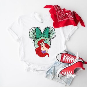 Puede incluir: Camiseta blanca con un gr&aacute;fico de Ariel de La Sirenita, un lazo verde con lunares y orejas de rat&oacute;n. Zapatillas rojas, pantalones cortos de mezclilla y una bandana roja completan el atuendo informal. Un dise&ntilde;o divertido y colorido.