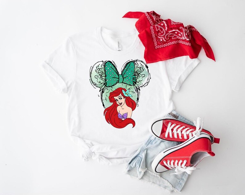 Puede incluir: Camiseta blanca con un gr&aacute;fico de Ariel de La Sirenita, un lazo verde con lunares y orejas de rat&oacute;n. Zapatillas rojas, pantalones cortos de mezclilla y una bandana roja completan el atuendo informal. Un dise&ntilde;o divertido y colorido.
