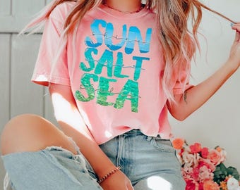 Könnte beinhalten: Ein korallenfarbenes Kurzarm-T-Shirt mit dem Schriftzug "SUN SALT SEA" in einem Farbverlauf von Blau zu Grün. Die Buchstaben haben ein strukturiertes, wasserähnliches Muster. Das T-Shirt wird mit einer hellblauen Jeans kombiniert.
