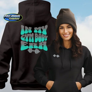 May include: A black hooded sweatshirt featuring a teal and white gradient graphic on the back that reads IN MY GYM BODY ERA with a dumbbell icon. The front of the hoodie displays a small dumbbell graphic on the chest. A black beanie is also shown.