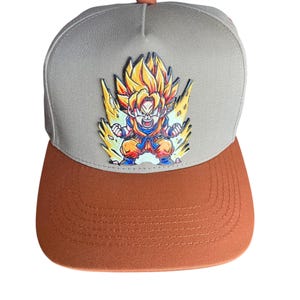 May include: A gray and orange baseball cap featuring a colorful graphic of a character with spiky yellow hair and blue clothing. The cap has a flat brim and a button on top. The design is from the Dragon Ball Z anime series.