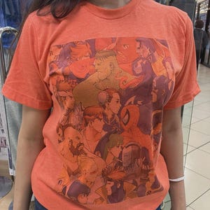 Könnte beinhalten: Orangefarbenes T-Shirt mit Rundhalsausschnitt und einer Collage von Comicfiguren. Das Design umfasst Hulk, Spider-Man und Captain America. Das T-Shirt besteht aus einem weichen Material und eignet sich für den Freizeitgebrauch. Das T-Shirt hat eine leuchtende orange Farbe.