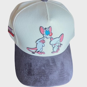 Puede incluir: Una gorra de béisbol con un panel frontal blanco que presenta un gráfico de los personajes de dibujos animados Pinky y Cerebro. La gorra tiene una visera y paneles laterales de aspecto de ante gris oscuro. El texto es visible en el lateral.