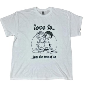 May include: White t-shirt with a black graphic of two cartoon figures embracing, sitting on a bench. The text "love is..." is above the image, and "...just the two of us" is below.