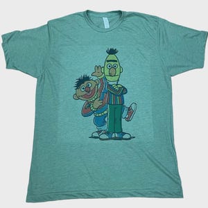 Op de afbeelding: Lichtgroen t-shirt met een kleurrijke afbeelding van Bert en Ernie van Sesamstraat. Bert draagt een gestreept shirt en groene broek, terwijl Ernie een blauwe en rode outfit heeft. Het shirt is gemaakt van zacht materiaal.