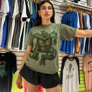 May include: An olive green t-shirt featuring a graphic of a Teenage Mutant Ninja Turtle. The shirt is paired with black shorts. Other t-shirts in various colors hang on a rack in the background.