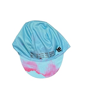 May include: A light blue cycling cap with a tie-dye pink and blue design on the brim. The cap has the text "Don't ride modes, make waves" printed on it in black. A black logo is on the side.
