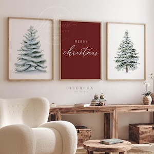 Può includere: Tre stampe artistiche da parete incorniciate che raffigurano due alberi sempreverdi innevati in stile acquerello su sfondo bianco e una stampa centrale rosso scuro con il testo bianco Merry Christmas in corsivo, in cornici di legno chiaro.