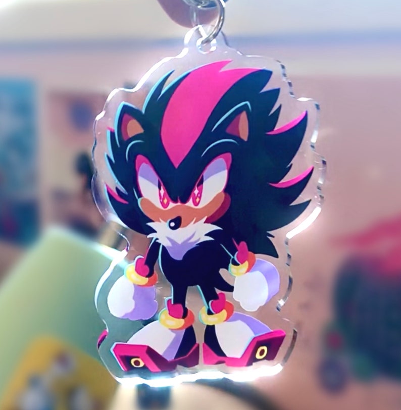 May include: A black and pink anime character with red eyes, a white muzzle, and gold bracelets. The character is standing on a clear acrylic keychain.