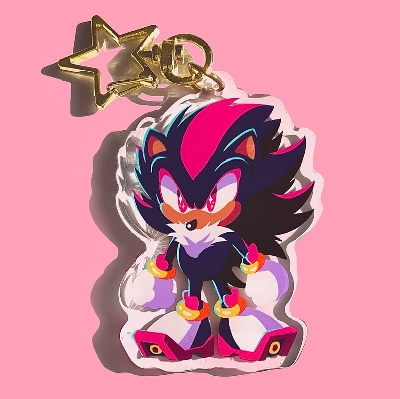 May include: A black and pink Shadow the Hedgehog keychain with a gold star charm. The keychain is made of acrylic and features a detailed illustration of Shadow with his signature red eyes and black quills.