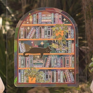 May include: A holographic sticker featuring a bookshelf design with a black cat lounging on the shelves. The shelves are filled with books and potted plants. The sticker has a rainbow iridescent effect and is set against a blurred green background.