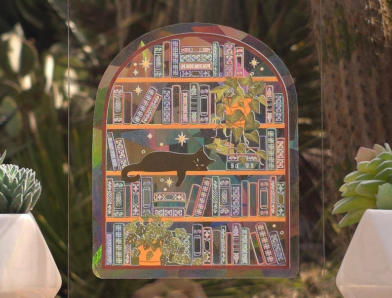 K&ouml;nnte beinhalten: Ein holografischer Aufkleber mit einem B&uuml;cherregal-Design, auf dem eine schwarze Katze liegt. Die Regale sind mit B&uuml;chern und Topfpflanzen gef&uuml;llt. Der Aufkleber hat einen regenbogenfarbenen, schillernden Effekt und ist vor einem verschwommenen gr&uuml;nen Hintergrund platziert.