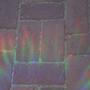 Pu&ograve; includere: Primo piano di un viale lastricato con un motivo arcobaleno creato dalla luce del sole che si riflette su un prisma. I mattoni sono di un colore marrone spento e l'arcobaleno &egrave; un mix vibrante di rosso, arancione, giallo, verde, blu, indaco e viola.