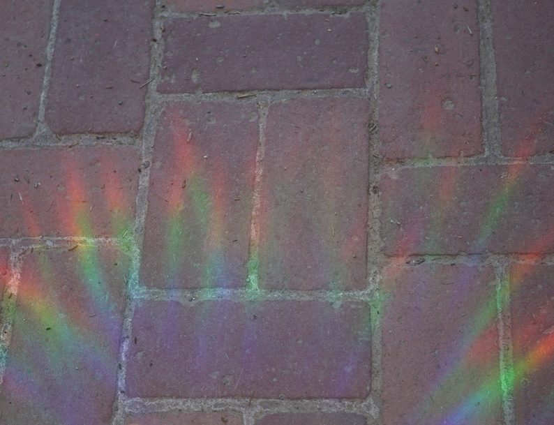 Pu&ograve; includere: Primo piano di un viale lastricato con un motivo arcobaleno creato dalla luce del sole che si riflette su un prisma. I mattoni sono di un colore marrone spento e l'arcobaleno &egrave; un mix vibrante di rosso, arancione, giallo, verde, blu, indaco e viola.