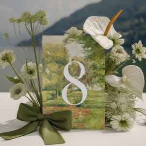 May include: A decorative card featuring a landscape painting with the number 8 in white. The card is adorned with white flowers, greenery, and a green satin bow. The background shows a lake and mountains.