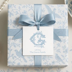Op de afbeelding: Een vierkante geschenkdoos verpakt in wit papier met een lichtblauw bloemen- en vogelpatroon. Een lichtblauw satijnen lint en strik sieren de bovenkant. Een wit label met een lichtblauwe sierlijke letter "G" en de tekst "with love, The Garcia Family" is bevestigd.