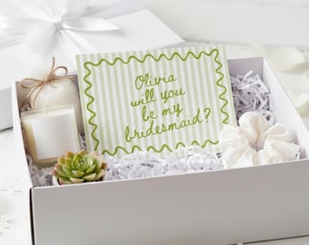 May include: A white gift box with a white bow on top, containing a candle, succulent, scrunchie, and a card that reads "Olivia will you be my bridesmaid?" The card has a green wavy border and green striped background.