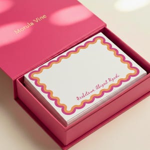 Può includere: Una scatola rosa con il testo "Morula Vine" sul coperchio, aperta per rivelare una pila di biglietti bianchi. Ogni biglietto ha un bordo smerlato arancione e rosa, con il nome "Reabetswe Abigail Kgadi" stampato.