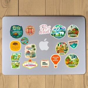 Puede incluir: Una colecci&oacute;n de pegatinas coloridas con tem&aacute;tica de viajes en un port&aacute;til. Las pegatinas presentan destinos como Alaska, Italia y Las Vegas. Los dise&ntilde;os incluyen un oso, un faro, un globo aerost&aacute;tico y texto como "Around the World".
