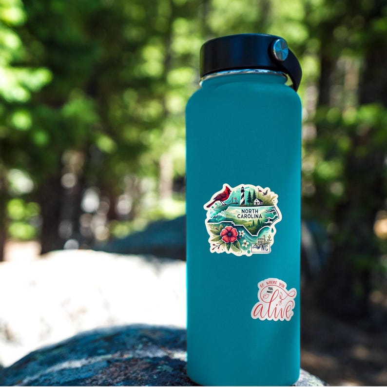 Puede incluir: Una botella de agua turquesa con tapa negra y asa plateada. La botella tiene dos pegatinas: una con un dise&ntilde;o de Carolina del Norte y un cardenal, y otra con el texto "Go where you feel most alive."