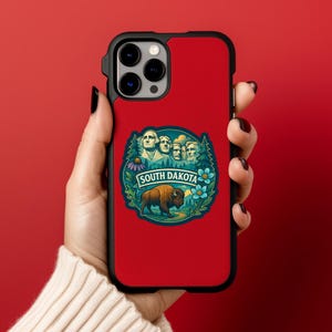 Puede incluir: Funda de tel&eacute;fono roja con borde negro. Presenta una colorida ilustraci&oacute;n del Monte Rushmore, un bisonte y las palabras "SOUTH DAKOTA". El dise&ntilde;o est&aacute; rodeado de &aacute;rboles y flores.