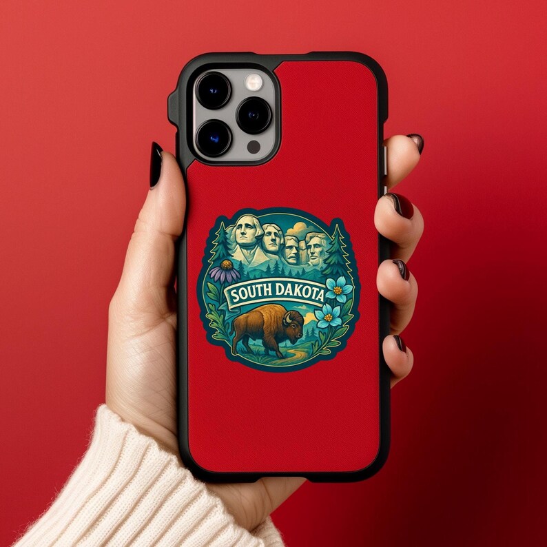 Puede incluir: Funda de tel&eacute;fono roja con borde negro. Presenta una colorida ilustraci&oacute;n del Monte Rushmore, un bisonte y las palabras "SOUTH DAKOTA". El dise&ntilde;o est&aacute; rodeado de &aacute;rboles y flores.