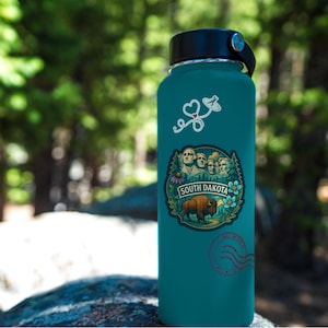 Puede incluir: Botella de agua aislada de color turquesa con tapa y asa negras. La botella presenta una pegatina con el texto "SOUTH DAKOTA" y una imagen del Monte Rushmore y un bisonte. Pegatinas adicionales incluyen un coraz&oacute;n y un sello.