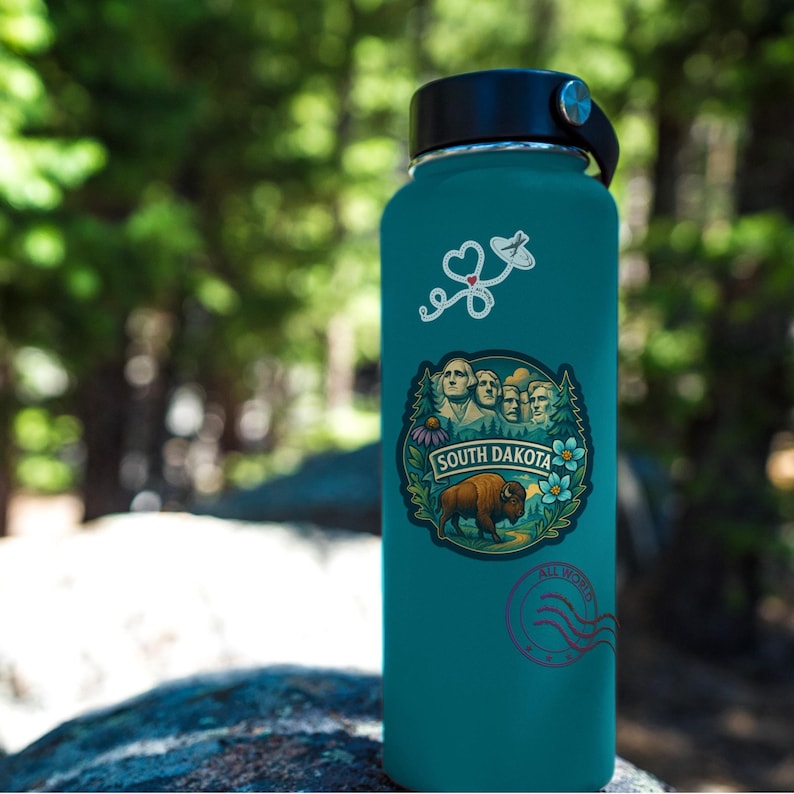 Puede incluir: Botella de agua aislada de color turquesa con tapa y asa negras. La botella presenta una pegatina con el texto "SOUTH DAKOTA" y una imagen del Monte Rushmore y un bisonte. Pegatinas adicionales incluyen un coraz&oacute;n y un sello.